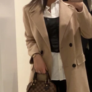 Beige kappa från Zara med knappar - Stilren beige kappa från Zara med dubbelknäppning och stora svarta knappar. Klassisk krage, raka fickor med lock och rak passform. Perfekt för dig som gillar en clean och tidlös look. Materialet är mjukt och känns lyxigt.