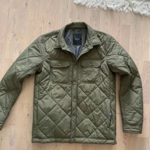 Olivgrön quiltad jacka Reserved S - Quiltad olivgrön jacka från Reserved i storlek S. Jackan har två bröstfickor med knappar, klassisk krage och snyggt rutmönster över hela ytan. Perfekt för dig som gillar stilren och funktionell design med en urban touch.