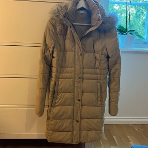 Beige dunjacka Tommy Hilfiger S - Snygg beige dunjacka från Tommy Hilfiger i storlek S. Jackan har en fluffig huva med fuskpäls, quiltad design och långa ärmar. Stängs med både dragkedja och knappar framtill. Perfekt för kalla dagar och har en stilren look.