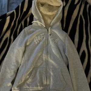 Grå hoodie från Abercrombie & Fitch - Mysig grå hoodie från Abercrombie & Fitch med broderad logga framtill. Tröjan har dragkedja, huva med snörning och är fodrad med fluffigt teddyfoder. Perfekt för kyliga dagar och har två fickor framtill.