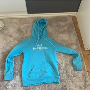 Turkos hoodie från Peak Performance - Snygg turkos hoodie från Peak Performance i storlek S. Tröjan har huva med snörning, magficka och vit logotyp på bröstet. Tillverkad i mjuk bomullsmix, perfekt för chill eller träning. Klassisk passform med långa ärmar och ribbade muddar.