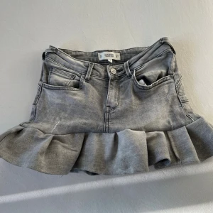 Grå handgjord jeanskjol med volang från Mango - Snygg grå jeanskjol handgjord av Mango jeans med volangkant nedtill. Kjolen har klassiska jeansdetaljer som fickor fram och bak, knapp och dragkedja. Perfekt för dig som gillar en cool och trendig stil med lite edge.