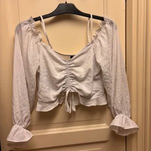 Vit topp med volang från H&M Stl 40 - Supersöt vit blus från H&M med långa puffärmar, volangdetaljer vid ärmslut och axlar samt snörning framtill. Blusen har v-ringning, tunna axelband och är croppad med lätt struktur i tyget. Perfekt för en trendig look! Jättefin!!