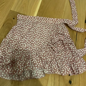 Rosa omlottkjol med volang och blommor - Supersöt kort omlottkjol i ljusrosa med vita blommor. Kjolen har volang längs nederkanten och knyts i midjan med ett brett band. Tillverkad i ett lätt och mjukt material som känns skönt mot huden. Perfekt för vår och sommar när du vill ha en lekfull och trendig look. En knapp har lossnat så priset är lägre