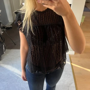 Svart blus med volangärmar och spets - Säljer en svart blus med transparent tyg och lodräta ränder. Blusen har volangdetaljer vid axlarna och är ärmlös. Den är luftig och har en lätt A-linjeform. 