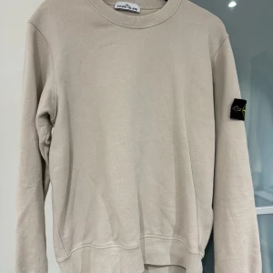 Beige sweatshirt från Stone Island - Säljer en stilren beige sweatshirt från Stone Island med klassisk rund hals och ikonisk logotyp-patch på vänster ärm. Tröjan är i mjuk bomull och har en relaxed passform med långa ärmar. Perfekt för dig som gillar clean och enkel streetwear.