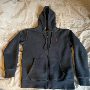 Marin blå hoodie från Polo Ralph Lauren M -  Polo Ralph Lauren med röd broderad logga på bröstet. Tröjan har dragkedja, snörning i huvan och stora fickor framtill. Tillverkad i mjuk bomull och perfekt för chill dagar. Klassisk och stilren design.
