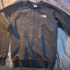 Svart sweatshirt från The North Face - Svart sweatshirt från The North Face med vit logga på bröstet och stort tryck på ryggen med texten 'Never Stop Exploring'. Tröjan har rund hals, långa ärmar och ribbade muddar. Perfekt för chill dagar eller när du vill ha en clean streetstyle.