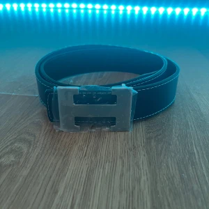 Blått skärp med H-spänne i silver - Snyggt blått bälte med ett stort H-format spänne i silverfärg. Bältet har vita kontrastsömmar längs kanterna och är gjort i skinn med en lätt texturerad yta. Perfekt för att lyfta vilken outfit som helst med en stilren detalj.