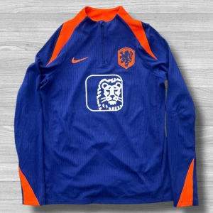 Holländsk långärmad Nike fotbollströja M - Säljer en blå långärmad fotbollströja från Nike med orange detaljer och KNVB-emblem. Tröjan har halv dragkedja, Dri-FIT ADV-material och vit lejonlogga på bröstet. Slim fit-modell, perfekt för träning eller match.