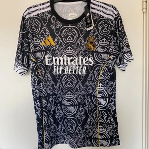 Real Madrid x Adidas svart matchtröja L - Snygg Real Madrid fotbollströja från Adidas i svart med vitt grafiskt mönster och guldiga detaljer. Slim fit-modell i lätt funktionsmaterial, klassiska tre ränder på axlarna och klubbmärke på bröstet. Perfekt för dig som vill sticka ut på planen.