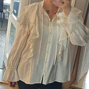 Vit volangskjorta med knappar - Skjorta med volanger längs axlar och bröst, knappar framtill och långa ärmar med volangdetalj vid ärmslut. Skjortan har en lätt glansig yta och en relaxed fit. 