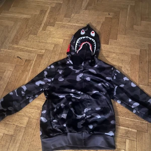 BAPE camo shark hoodie svart/grå - Säljer en ikonisk BAPE shark hoodie med svart och grått camo-mönster. Huvan har hajansikte med tänder och ögon, samt broderade bokstäver och tigertryck på sidorna. Jackan har dragkedja hela vägen upp.