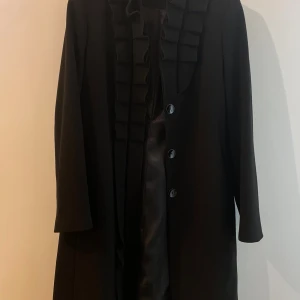 Svart ZeroPoint kappa - Stilren svart kappa från ZeroPoint, köpt från Turkiet. Snygga volangdetaljer längs kragen och knäppning framtill, samt en glansig foder, perfekt för en elegant look!