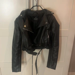Svart skinnjacka från Stradivarius XS - Säljer en svart skinnjacka från Stradivarius i storlek XS. Jackan har klassisk bikerstil med dragkedjor, knappar och bälte i midjan. Snygg croppad passform och coola detaljer som ger en edgy vibe. Perfekt att slänga över en hoodie eller t-shirt.
