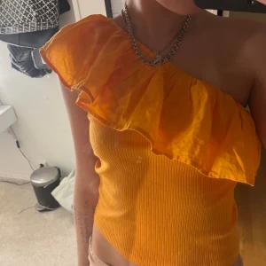 Orange oneshoulder topp med volang - Säljer en snygg orange oneshoulder topp med stor volang över axeln. Toppen är ribbad och har en tight passform som framhäver figuren. Perfekt för dig som vill sticka ut med färg och cool design.