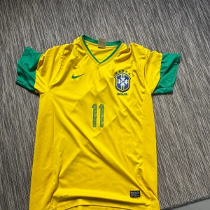 Brasilien Neymar 11 Nike fotbollströja - Gul Brasilien fotbollströja från Nike med gröna detaljer och nummer 11 på bröstet och ryggen. Neymar står tryckt på ryggen. Tröjan har korta ärmar, V-ringad krage och är tillverkad i lätt, ventilerande material. Perfekt för dig som älskar fotboll och Brasilien.