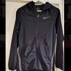 Svart Nike Dri-Fit träningsjacka XS - Svart Nike Dri-Fit träningsjacka med huva och hel dragkedja. Jackan har grå detaljer längs sidorna, två fickor framtill och är tillverkad i lätt och stretchig polyester. Perfekt för träning eller aktiva dagar. Klassisk Nike-logga på bröstet.