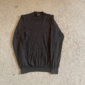 Mörkgrå rundhalsad tröja Oscar Jacobson - Snygg mörkgrå rundhalsad tröja från Oscar Jacobson i storlek S. Tröjan är tillverkad i Italien och har en klassisk, stilren design med ribbade muddar vid ärmslut och nederkant. Perfekt för dig som gillar enkel och tidlös stil.