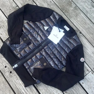 Svart quiltad cardigan jacka Moncler - Svart cardigan jacka från Moncler med quiltad, glansig front och stickade ärmar. Jackan har två fickor med knappstängning, rund hals och dragkedja framtill. Moncler-logga på ärmen. Perfekt för dig som gillar stilren och exklusiv look.