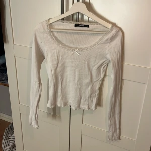 Vit ribbad topp med spets från Bikbok - Säljer en vit långärmad topp från Bikbok i ribbad bomull. Toppen har en söt spetskant runt halsen och en liten rosett framtill. Modellen är slim och har en lätt croppad passform med vågig nederkant. Perfekt för dig som gillar romantiska detaljer.