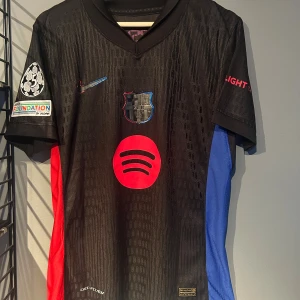 FC Barcelona Raphinha 11 Nike tröja S - Säljer en officiell FC Barcelona matchtröja från Nike med Raphinha 11 på ryggen. Tröjan är svart med röda och blå detaljer, korta ärmar och klubbmärke på bröstet. Materialet är lätt och andas, perfekt för fotboll. Spotify-logga och Champions League-patch på ärmen. Helt ny tröja med tagg. En replika
