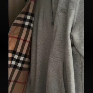 Grå hoodie från Burberry  - Säljer en grå hoodie från Burberry i storlek S med klassiskt rutigt foder i beige, svart, vitt och rött. Tröjan har snörning vid huvan, half zip och broderad logga på bröstet. Tillverkad i mjuk bomull, perfekt för dig som gillar stilrena detaljer och lyxig känsla.