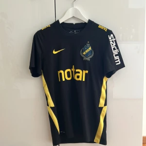 AIK svart fotbollströja Nike - AIK:s officiella matchtröja, 130 års jubileum, svart med gula detaljer och klubbmärke på bröstet. Tillverkad i lätt och ventilerande material, med tryckta sponsorer och gula inslag längs sidorna. Perfekt för dig som vill visa ditt stöd på läktaren eller planen.