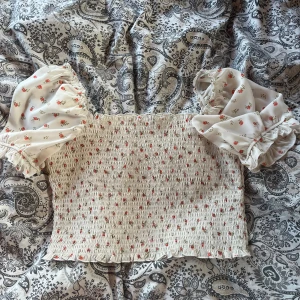 Blommig blus med puffärm från NA-KD - Supersöt blommig blus från NA-KD i vitt med små röda blommor och gröna blad. Toppen har smockad kropp, volangkant och puffiga korta ärmar med resår och volang. Har två små hål vid ärmarnas resår.Använd någon gång, säljer för den är lite liten på mig och lite kort för min smak