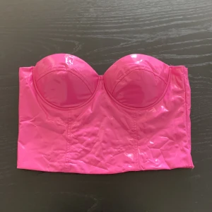 Rosa korsettopp från NA-KD - Säljer en glansig rosa korsettopp från NA-KD i storlek 38. Toppen är axelbandslös med vadderade kupor och dragkedja bak. Materialet är syntet och har en cool, nästan lackad finish som verkligen sticker ut. Liten fläck bak.