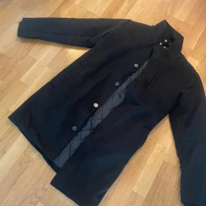 Svart rock med quiltad insida - Snygg svart rock med hög krage och knappar framtill. Insidan har ett quiltat mönster som ger extra värme och stil. Jackan har två sidofickor och en stilren, rak passform som funkar till många olika outfits.