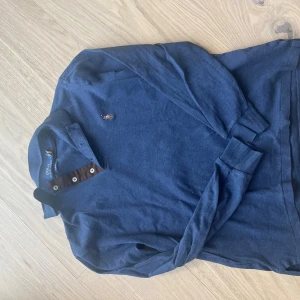 Blå pikétröja från Polo Ralph Lauren - Snygg marinblå pikétröja från Polo Ralph Lauren i slim fit-modell. Tröjan har långa ärmar, klassisk krage och knappar framtill. Ikoniska broderade loggan på bröstet. Perfekt för dig som gillar stilrena och tidlösa plagg. Nypris 1000-1500