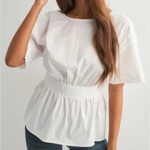 Vit blus med öppen rygg och rosett - Säljer en vit blus med korta ärmar och bred midjedetalj. Blusen har en snygg öppen rygg med stor rosett som knyts baktill. Materialet är mjukt och lätt bomull, perfekt för en fräsch och stilren look. Passformen är normal och blusen har en rund halsringning framtill.