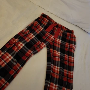 Röd-rutiga mjukisbyxor med snörning - Supermysiga mjukisbyxor i röd, svart och vit rutig design med bred resår i midjan och röd snörning. Perfekta för chill hemma eller myskvällar. Tillverkade i mjukt bomullsmaterial som känns skönt mot huden.