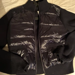 Svart pufferjacka från Gina Tricot - Svart pufferjacka från Gina Tricot med stickade ärmar och rygg. Jackan har ribbad krage, muddar och två fickor framtill. Framsidan är quiltad och glansig medan resten är stickad, vilket ger en cool kontrast. Dragkedja hela vägen fram.