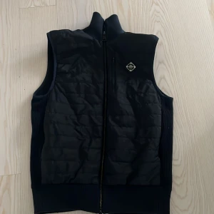  Väst J.Lindeberg - Svart dunväst från J.Lindeberg med quiltad framsida, ribbade sidor och hög krage. Västen har dragkedja framtill och en snygg logotyp på bröstet. Perfekt för lager-på-lager och stilren look.