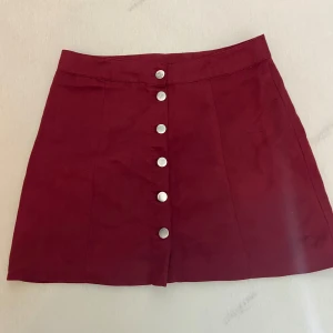 Vinröd mockakjol med knappar - Vinröd kortkjol i mjuk mocka-look med silvriga knappar framtill. Kjolen har en A-linjeformad passform och är perfekt för dig som vill ha en stilren men ändå trendig vibe. Enkel att matcha med både stickade tröjor och t-shirts.