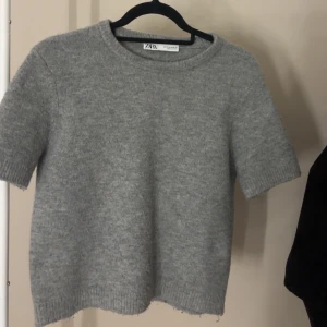 Grå stickad kortärmad tröja från Zara - Säljer en enkel och stilren grå stickad tröja från Zara i storlek M. Tröjan har korta ärmar, rund hals och ribbade kanter vid ärmslut och nederkant.
