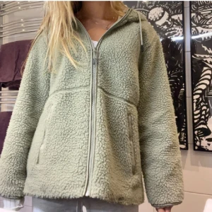 Grön teddyjacka med huva och dragkedja - Mysig ljusgrön teddyjacka med huva och dragsko, hel dragkedja framtill och två fickor. Jackan har en loose passform och är tillverkad i fluffigt fleece-material som håller dig varm. Perfekt för kyliga dagar när du vill vara både bekväm och snygg.