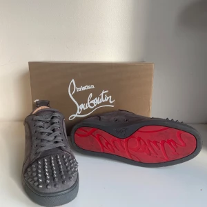 Gråa Christian Louboutin sneakers med nitar - Säljer ett par gråa sneakers från Christian Louboutin med ikonisk röd sula och coola silverfärgade nitar på tån. Skorna är i mocka och har snörning framtill. Perfekta för dig som vill sticka ut med en lyxig och edgy stil.