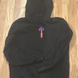 Svart Trapstar hoodie - Svart hoodie från Trapstar med stor färgglad T-logga och tryck i blått, rött och gult på både bröstet och ryggen. Klassisk huva med snörning och ribbade muddar. Perfekt för dig som gillar streetwear och vill sticka ut med snygga detaljer.