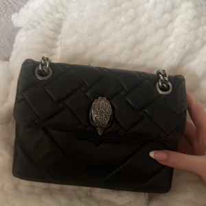 Svart quiltad axelväska Kurt Geiger - Snygg svart axelväska från Kurt Geiger i äkta skinn med quiltad design och silverfärgade kedjedetaljer. Väskan har ett unikt spänne framtill och diskret logga på baksidan. Perfekt storlek för det viktigaste den lilla modellen 