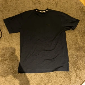 Svart BOSS t-shirt med camo-tryck - Svart t-shirt från BOSS i mjuk bomull med klassisk rund hals och korta ärmar. Framsidan har en diskret BOSS-logga, medan ryggen har ett stort tryck med grönt och blått camouflage-mönster och grafiska ansikten. Priset går att diskutera!