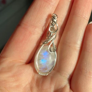 Berlock med regnbågsmånsten och silvertråd - Unik berlock med en oval regnbågsmånsten som skimrar i blått och vitt. Stenen är omsorgsfullt infattad i snirklig silvertråd som ger en bohemisk och magisk vibe. Perfekt för dig som älskar kristaller och vill ha något som sticker ut.