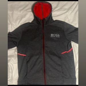 Grå Hugo Boss hoodie med röda detaljer - Snygg grå hoodie från Hugo Boss med röda detaljer och dragkedja framtill. Hoodien har röd insida i huvan, vita logotypdetaljer på bröstet och praktiska fickor. Perfekt för dig som gillar stilren streetwear med en twist.