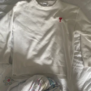 Vit stickad tröja från Ami - Vit stickad tröja från Ami med rund hals och ribbade muddar. På bröstet finns Ami:s ikoniska röda hjärta med A-broderi. Tröjan har lång ärm och är gjord i mjukt material som känns skönt mot huden. Perfekt för dig som gillar clean och stilren design.