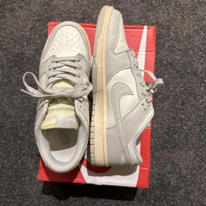 Nike Dunk Low i ljusgrått skinn - Nike Dunk Low sneakers i ljusgrått skinn med klassisk vit sula och matchande grå snörning. Snygg minimalistisk look med perforerad tå och ikonisk Swoosh på sidan. Perfekta för dig som gillar stilrena och tidlösa sneakers.