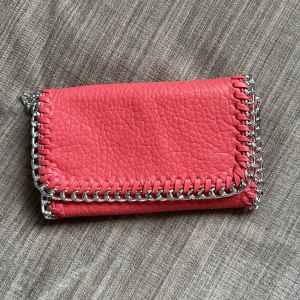 Rosa kuvertväska med kedjedetaljer från Tiamo - Snygg rosa kuvertväska från Tiamo i skinnimitation med silverfärgad kedja runt kanterna. Väskan har magnetknäppning, innerfack med dragkedja och avtagbar axelrem i metall. Perfekt för dig som vill sticka ut med en cool accessoar.