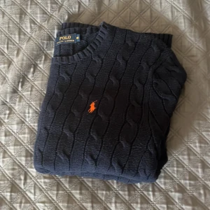Mörkblå kabelstickad tröja Polo Ralph Lauren - Säljer en mörkblå kabelstickad tröja från Polo Ralph Lauren i storlek M. Tröjan har rund halsringning, orange broderad logga på bröstet och klassisk ribbad kant. Perfekt för dig som gillar stilren och tidlös design.