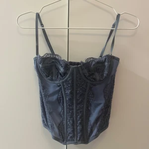 Blå spetskorsett - Supersnygg blå korsett med spetsdetaljer och smala justerbara axelband. Toppen har markerade sömmar och hakknäppning bak. Köpt på plick.
